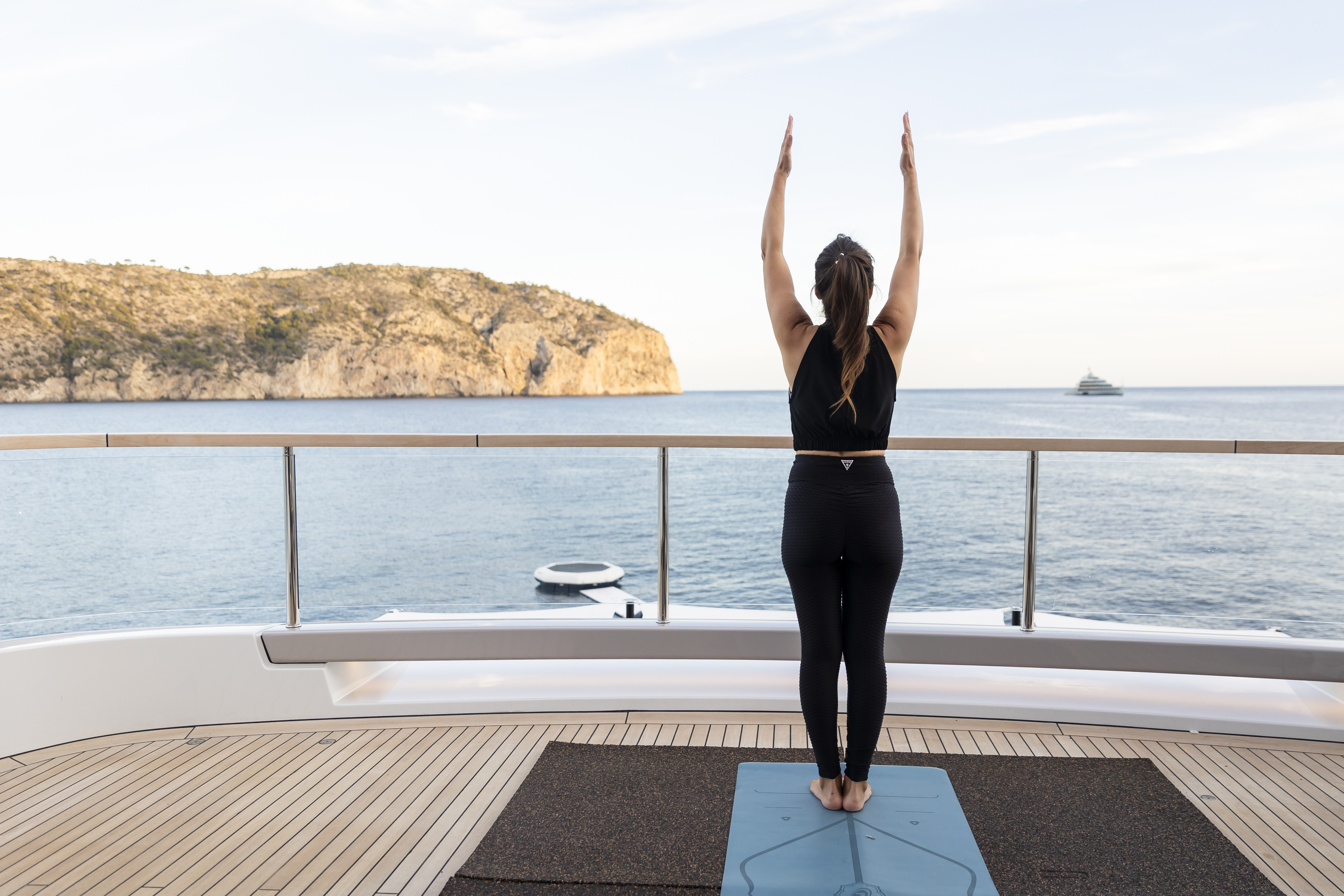 A woman practicing yoga on a deck overlooking the ocean, with her arms raised towards the sky and a scenic cliff in the background.